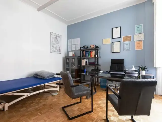 Uno studio medico con una scrivania, due sedie, un lettino per le visite, librerie e certificati incorniciati alla parete presso lo studio Fisioprati Roma. {{brizy_dc_image_alt entityId=